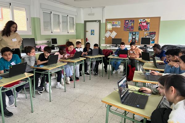 Niños y niñas trabajan con ordenadores portátiles en un aula, acompañados por una formadora, dentro del programa Menudos Techies.